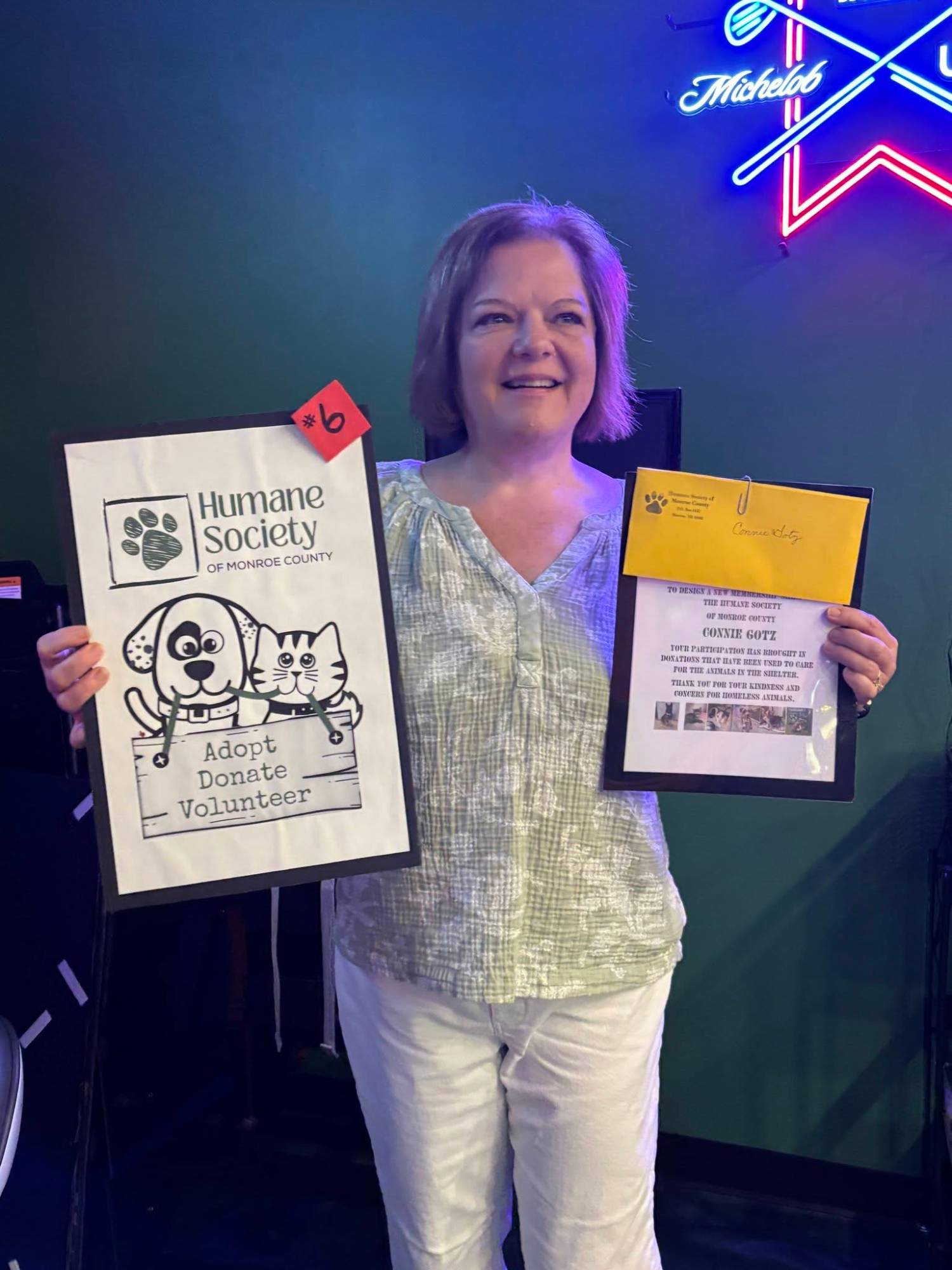 Connie Gotz holding her first-place design, entry #6, a hand-drawn Humane Society of Monroe County dog and cat with the words Adopt, Donate, Volunteer.
